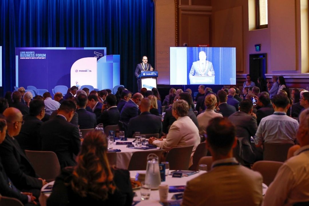 A speaker addresses a large audience in a conference room, with blue curtains and two large screens displaying the presentation. Attendees are seated at round tables.