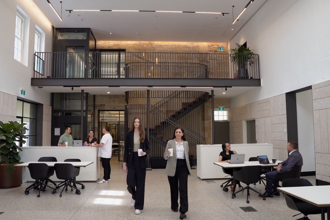 A modern office lobby with people walking and sitting. Glass walls, desks, and a staircase give it an open feel. Bright lighting creates a professional atmosphere.
