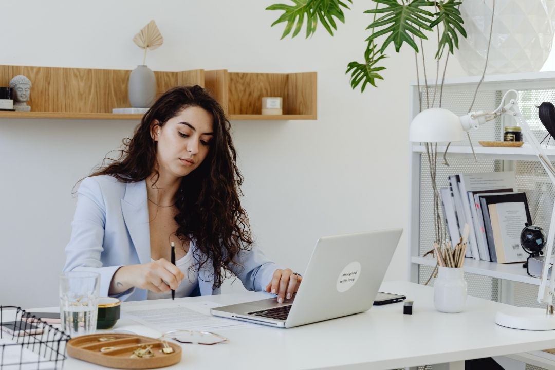 Woman in a home office, focused on writing in a notebook, a laptop open beside her. The setting is bright and modern, with shelves and plants.