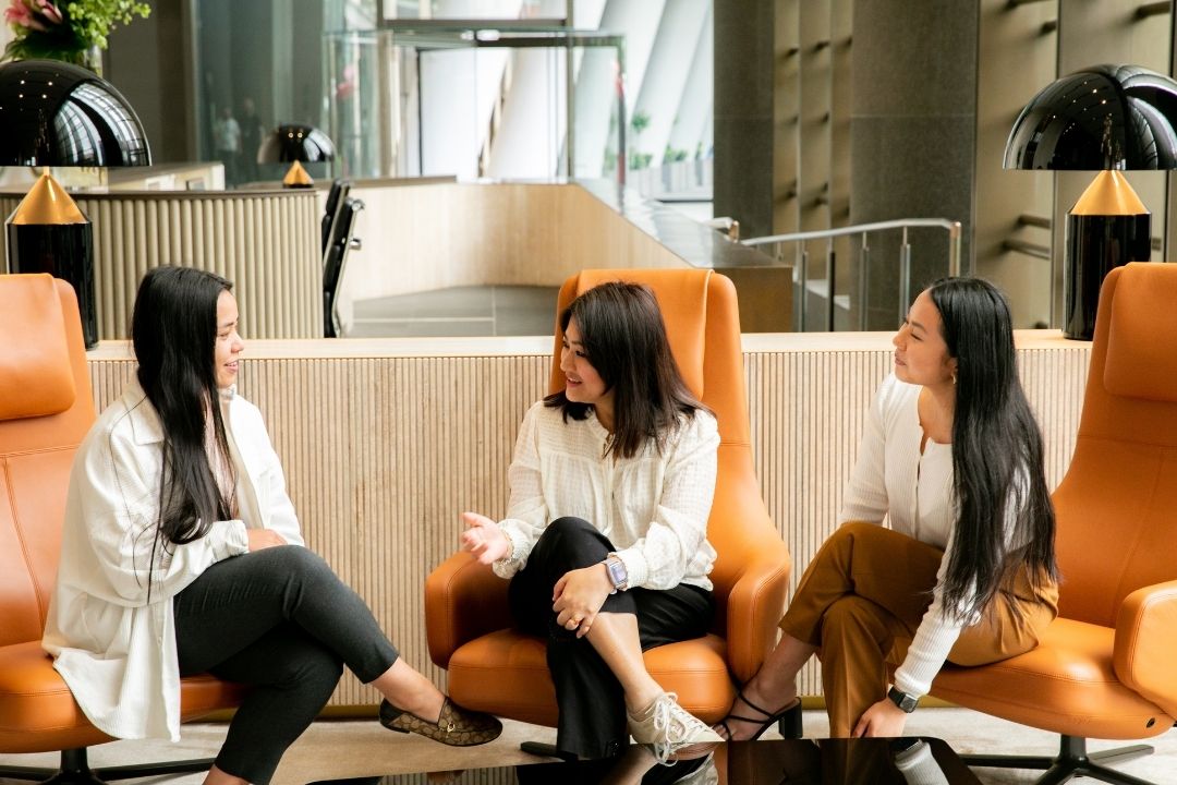 Three women in business attire sit on orange chairs, engaging in a lively discussion. The setting is a modern office with large windows and a bright atmosphere.
