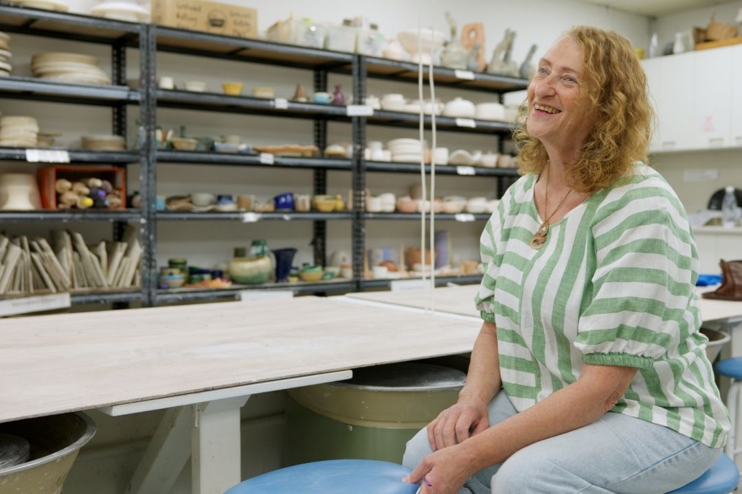 Woman smiling in a ceramics studio, wearing a striped shirt. Shelves lined with pottery and worktables surround her, conveying a creative ambiance.