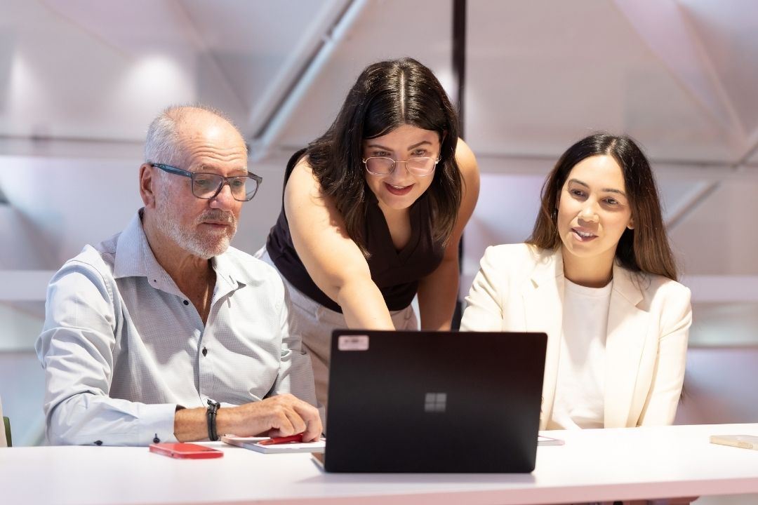A woman stands, pointing at a laptop screen, engaging with a seated man and woman. They are smiling, in a bright office setting, suggesting teamwork.