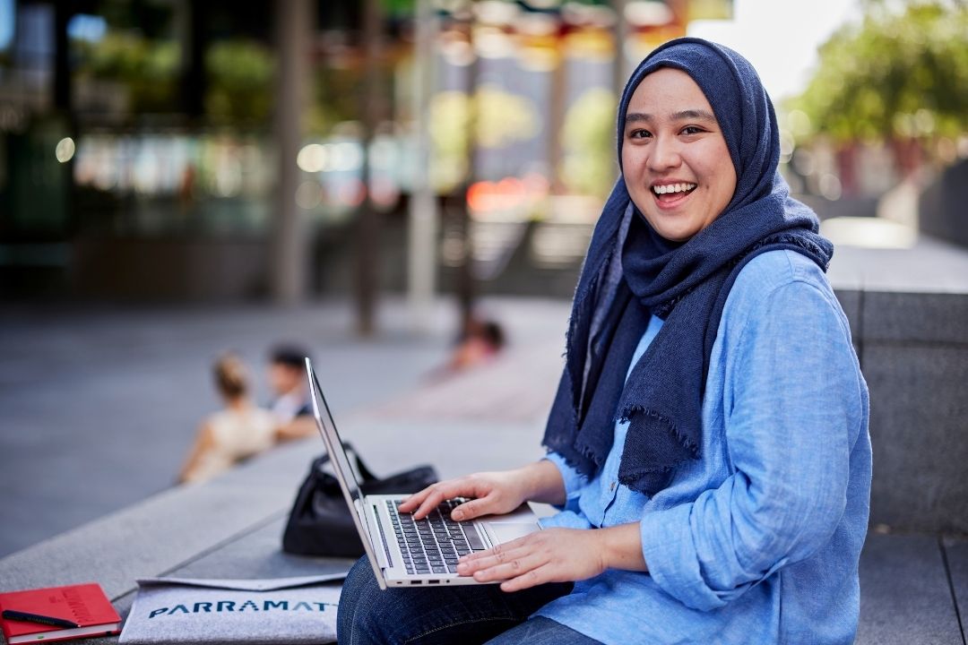 A woman wearing a blue hijab and shirt smiles while using a laptop outdoors on a sunny day. The background is a blurred urban setting with trees.