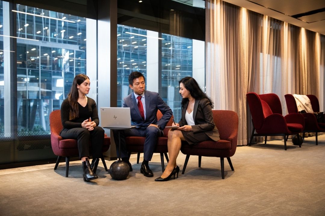 Three professionals in formal attire engage in a discussion around a laptop in a modern office lounge with large windows, exuding a collaborative atmosphere.