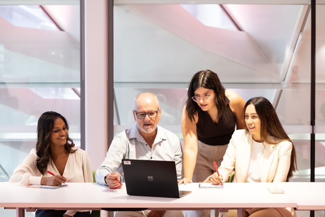 A diverse group of four professionals collaborate around a laptop at a bright office table, conveying teamwork and cooperation.