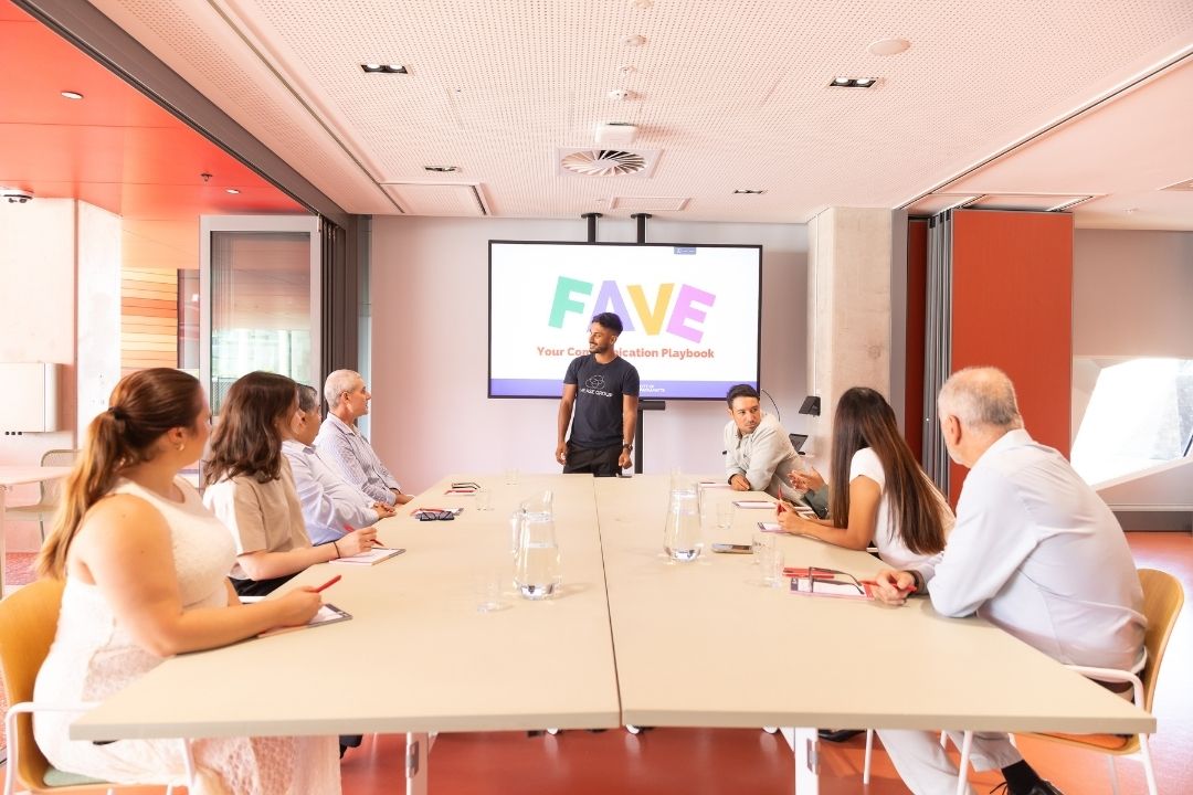 A diverse group in a bright conference room listens to a presenter in front of a screen. The atmosphere is focused and professional.