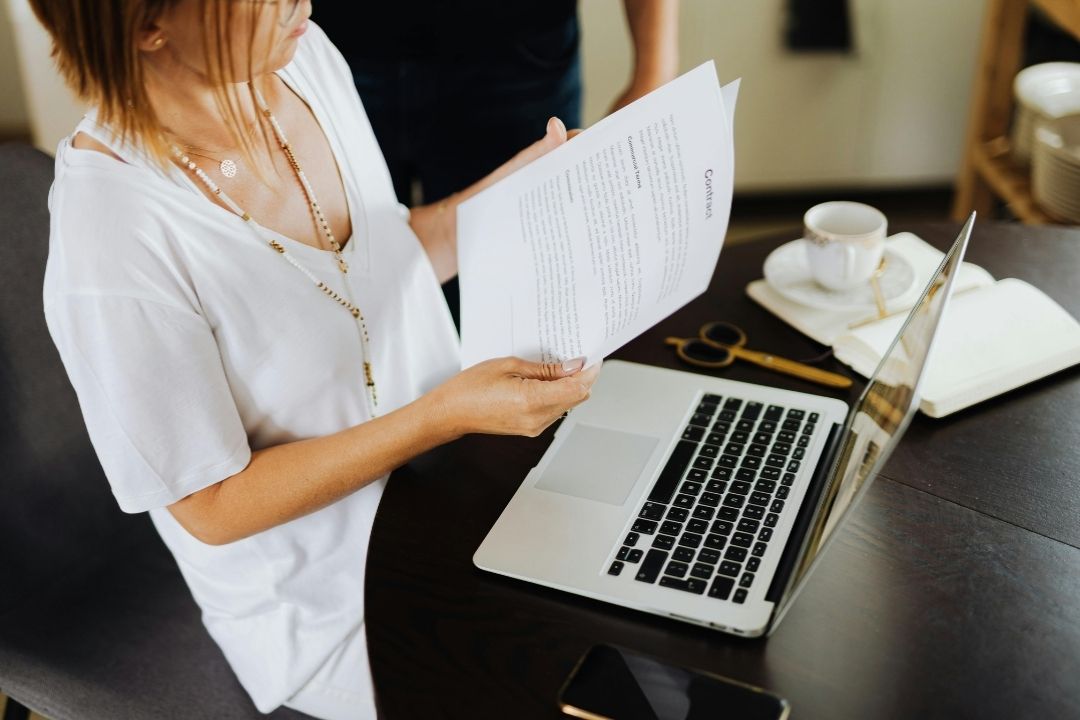 A person stands at a desk holding papers, with a laptop open and a coffee cup nearby. The scene conveys a busy and focused work environment.