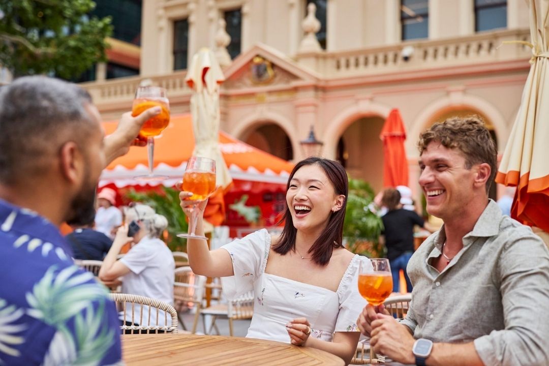 A group of three people enjoy drinks and laughter at an outdoor café with vibrant orange umbrellas, set against a backdrop of historic architecture.