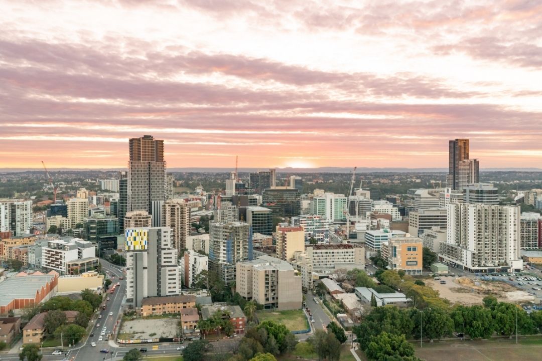 Aerial view of a city skyline at sunset, featuring tall skyscrapers and smaller buildings under a pink and orange sky, conveying a serene urban atmosphere.