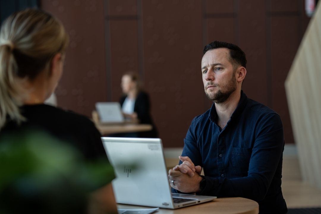 A man sits at a table with a laptop, attentively listening to a woman facing him. Another person is working at a laptop in the background. The setting is calm and professional.