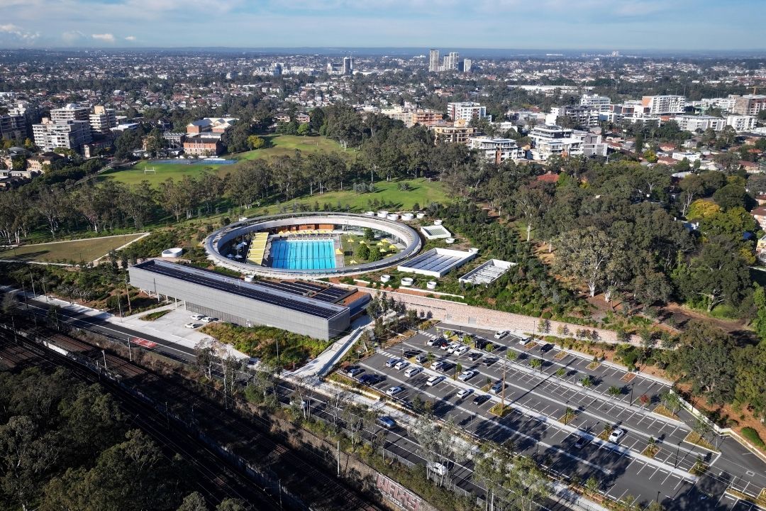 Aerial view of a modern sports stadium with blue seating, surrounded by trees and cityscape. Adjacent is a parking area with numerous cars.