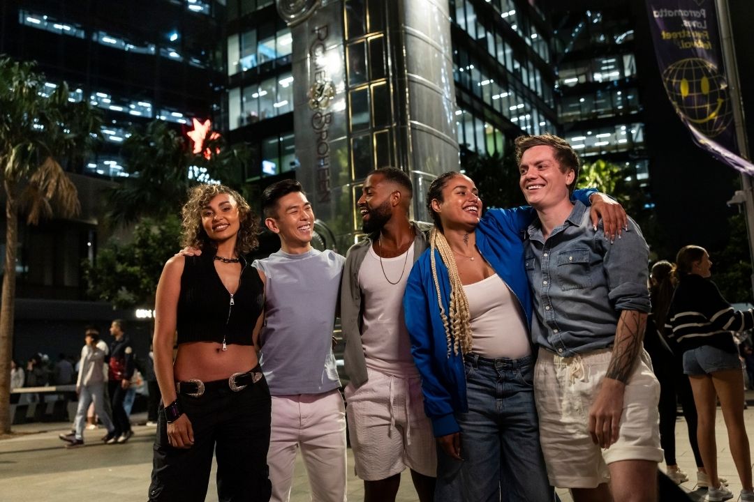 A group of five people stand closely together, smiling warmly, with city skyscrapers lit up behind them at night, creating a lively and joyful atmosphere.
