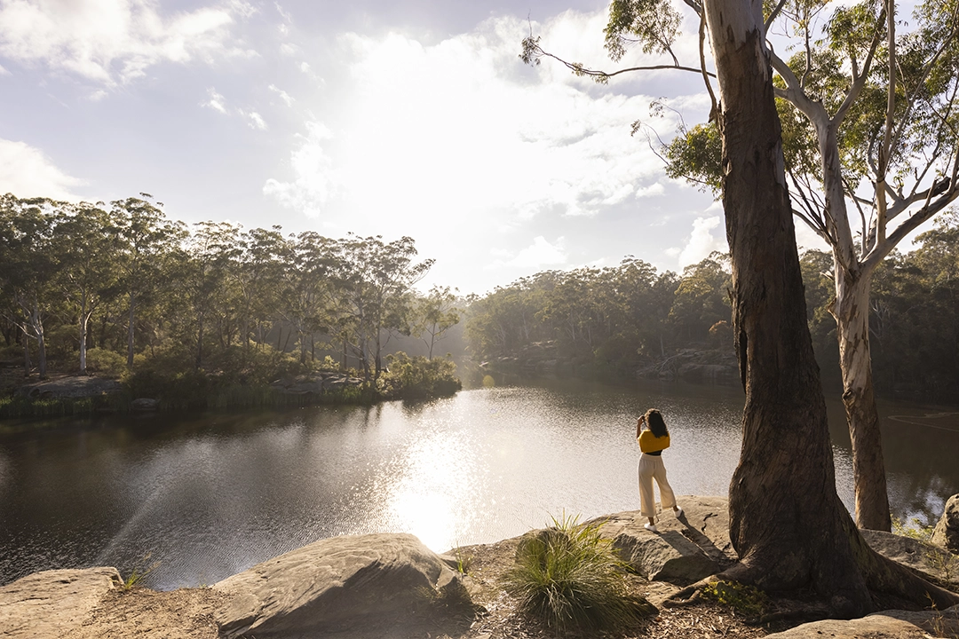 A person in a yellow top stands by a tranquil lake, surrounded by trees under a bright, cloudy sky.