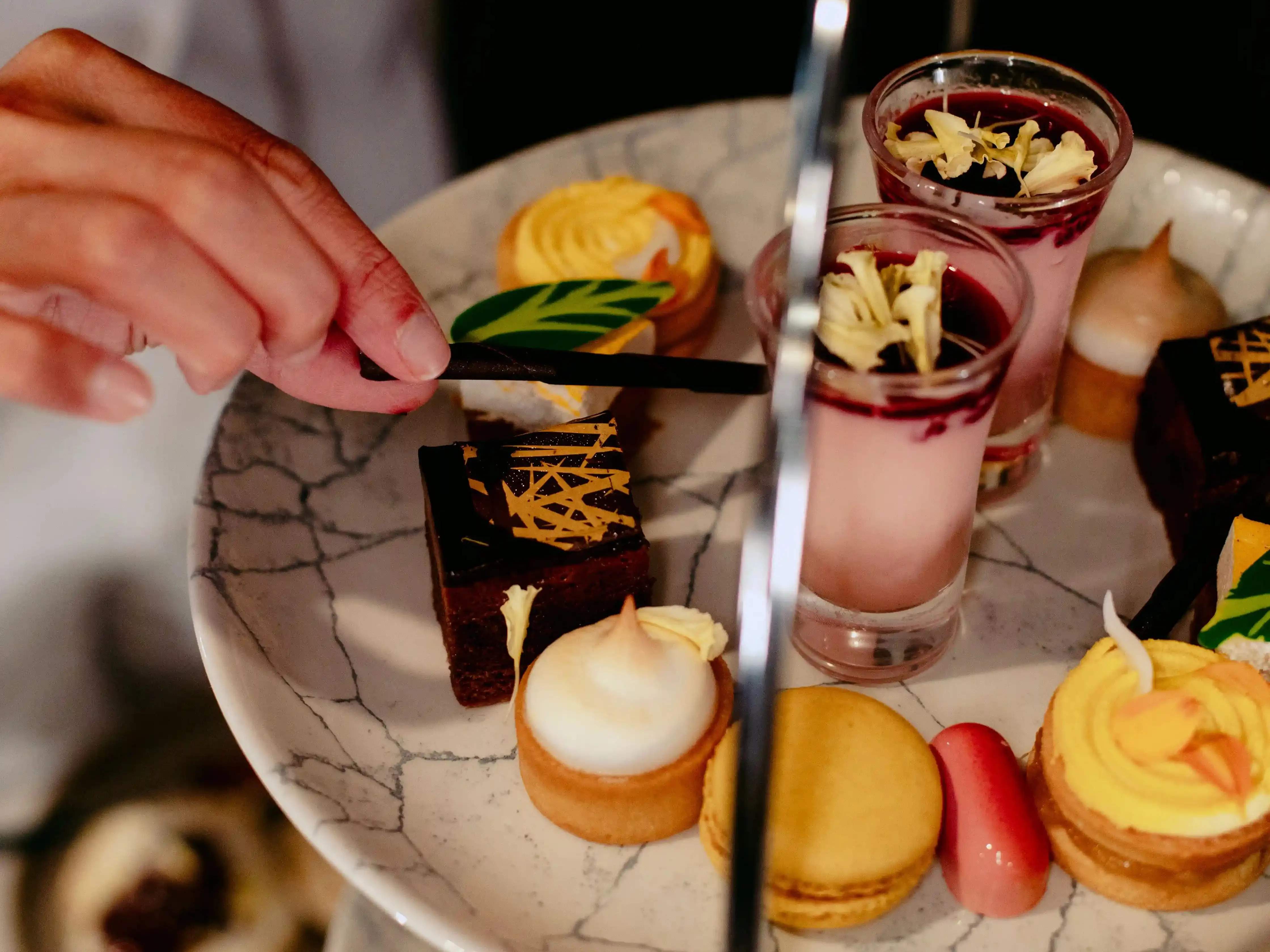 A hand arranges an elegant tiered tray with assorted desserts, including macarons, cakes, and mousse