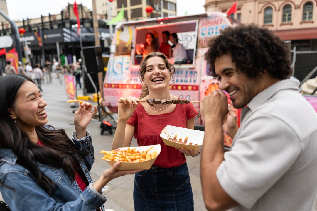 Three friends enjoy food from a street vendor, laughing together. The backdrop shows a vibrant street with colorful food trucks and people.