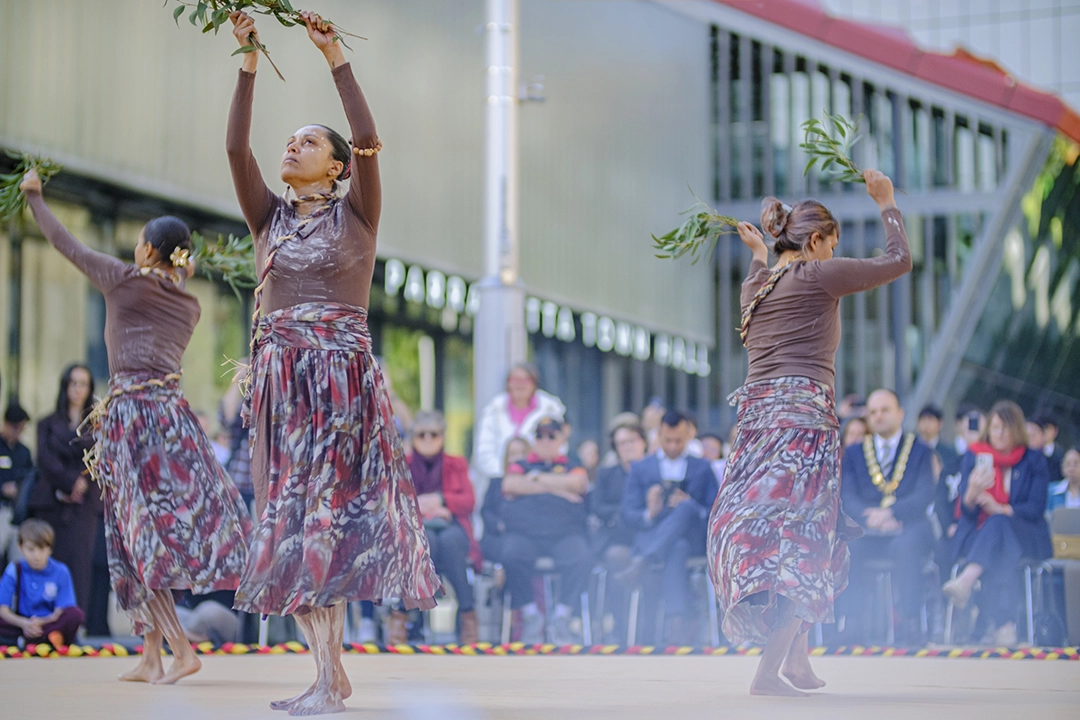 Three dancers in floral dresses perform gracefully outdoors, surrounded by a crowd. Mist adds a dreamy effect, conveying joy and celebration.