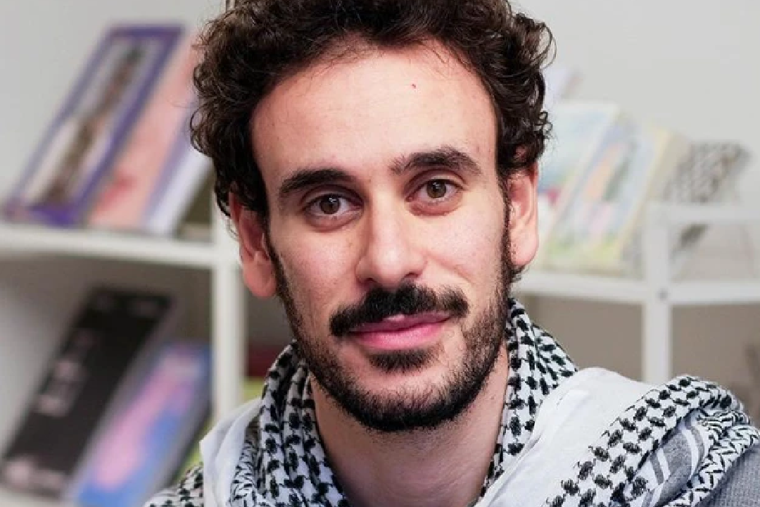 Young woman with long dark hair leans against a blue wall, looking thoughtfully at the camera. She wears a necklace and casual outfit, conveying a calm mood.