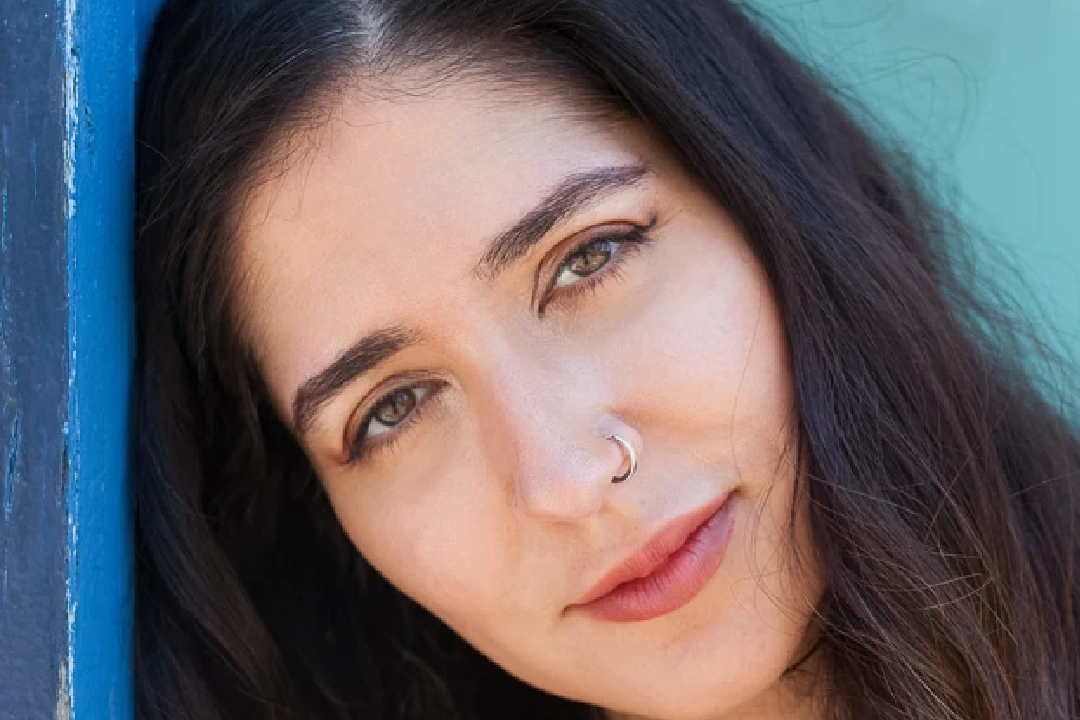 Young woman with long dark hair leans against a blue wall, looking thoughtfully at the camera. She wears a necklace and casual outfit, conveying a calm mood.