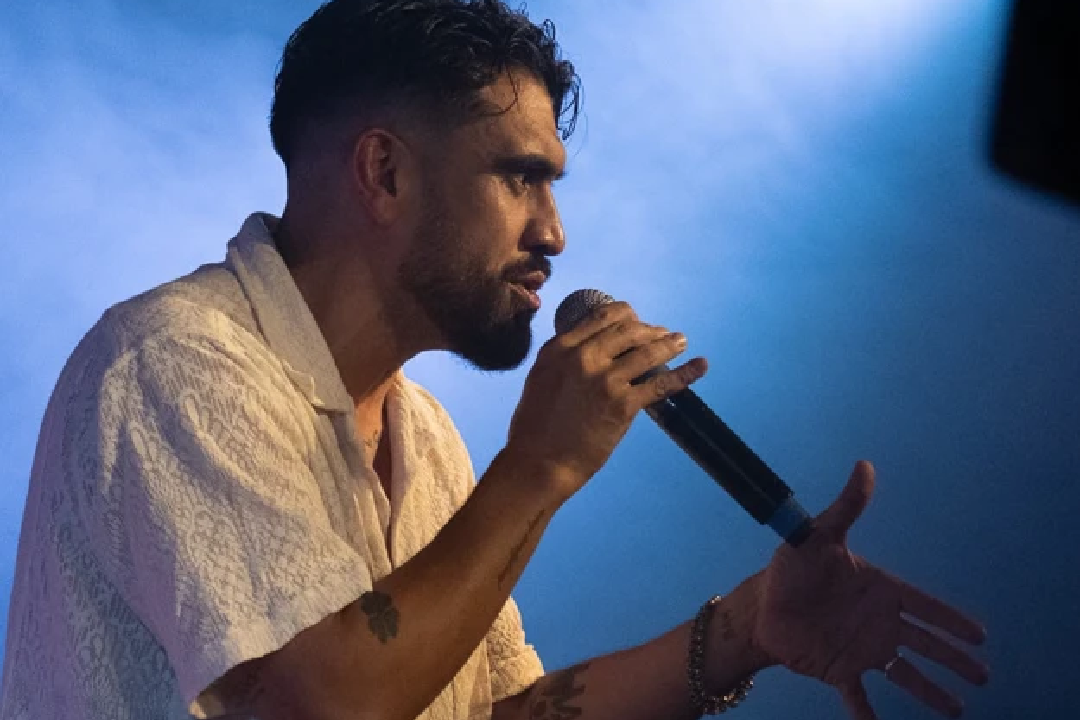 A man passionately performs on stage under blue lights, holding a microphone. He wears a light-colored shirt and gestures with his free hand, exuding energy.