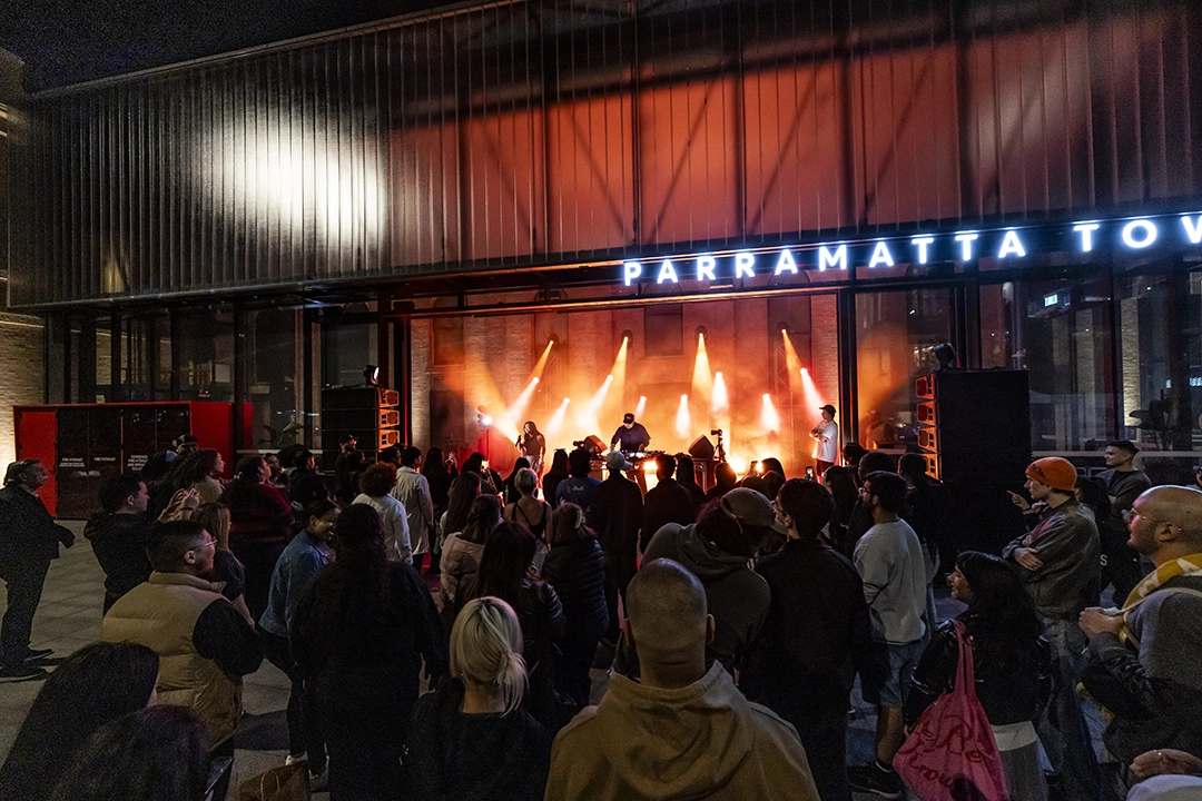 A lively crowd gathers at night in front of an illuminated Parramatta Town Hall, enjoying an engaging street performance under warm, glowing lights.