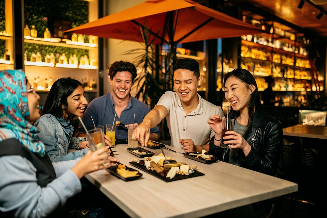 Five people sit around a table in a cozy restaurant, sharing laughter and desserts. Warm lighting and shelves of bottles create an inviting atmosphere.