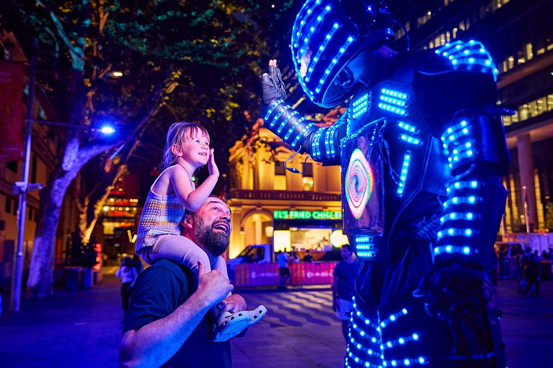 A child, sitting on a person's shoulders, reaches out to a performer in a dazzling blue LED suit at a lively night festival, evoking awe and wonder.