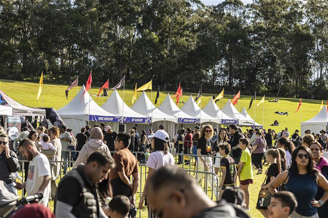 A lively outdoor festival scene with a crowd enjoying various stalls under white tents on a sunny day. Green trees and a bright grassy hill serve as a backdrop.