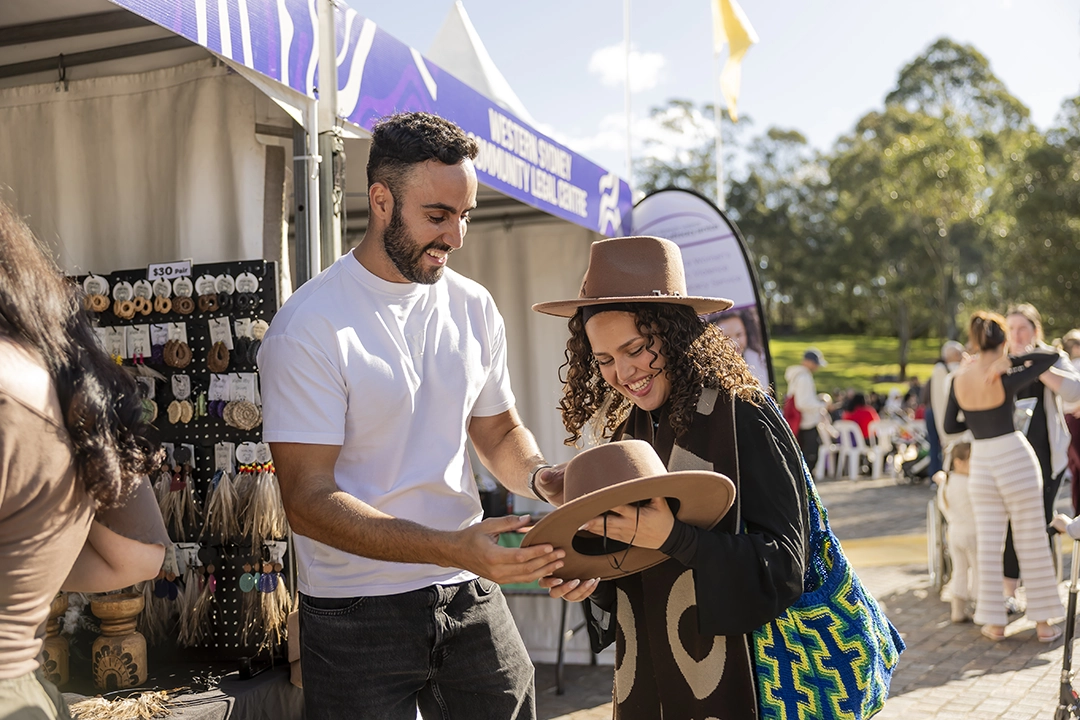 A woman and a man smile while looking at items at an outdoor market. She wears a wide-brimmed hat and holds a striped bag. Sunlight filters through trees.