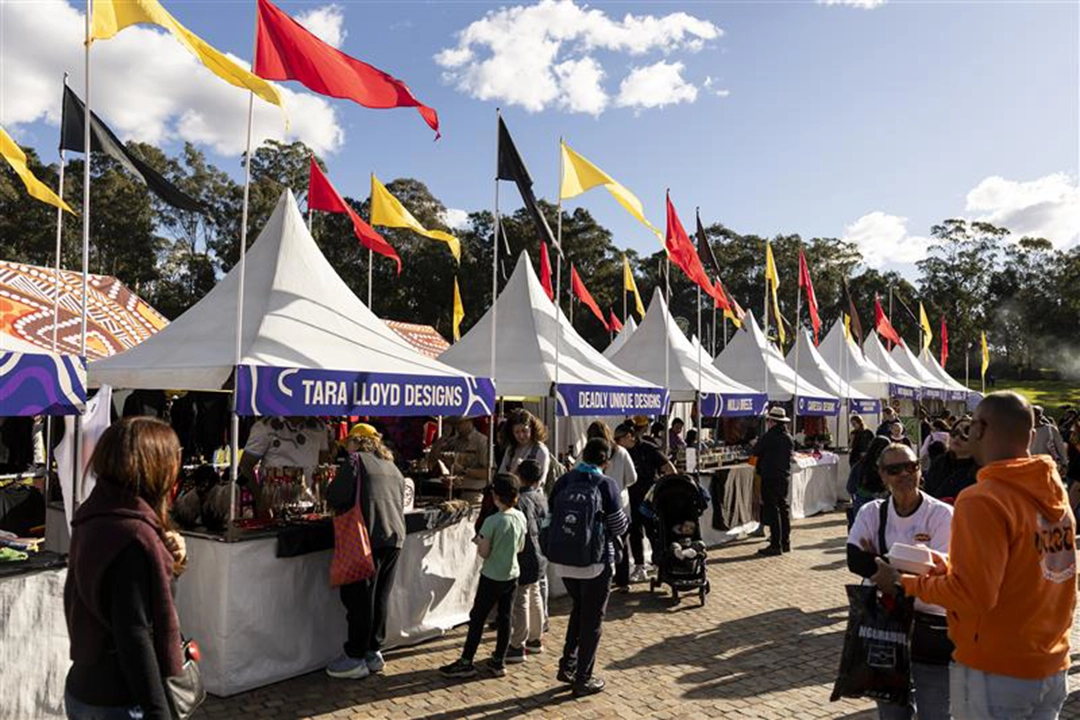 A bustling outdoor market with white tents lined up, international flags waving above. People are engaging with vendors under a sunny, cloud-dotted sky.