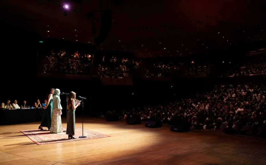 A singer performs on a warmly lit stage, facing a large, attentive audience in an auditorium. The atmosphere is elegant and intimate.