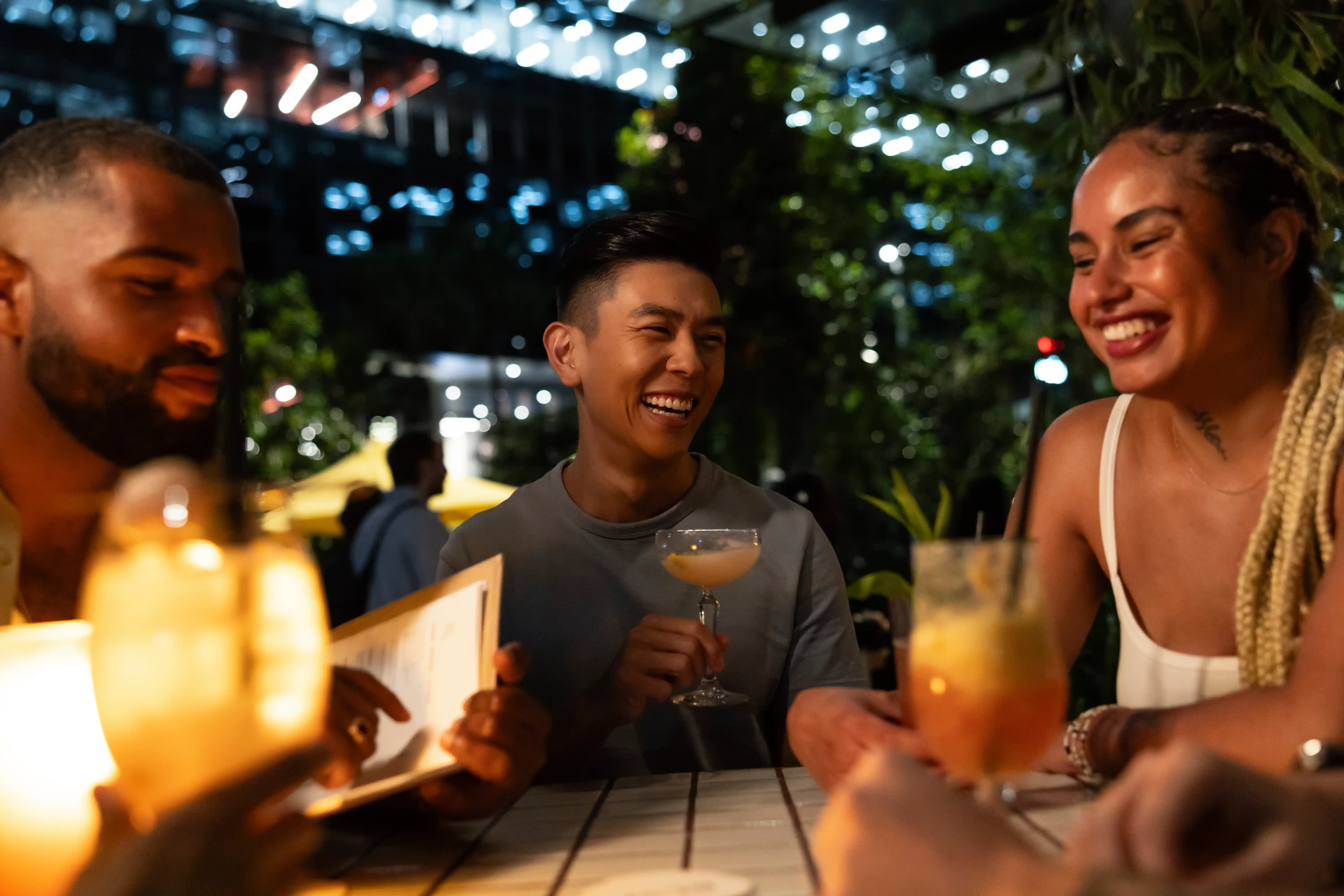 A group of three friends enjoy drinks at an outdoor table in a warmly lit urban setting.