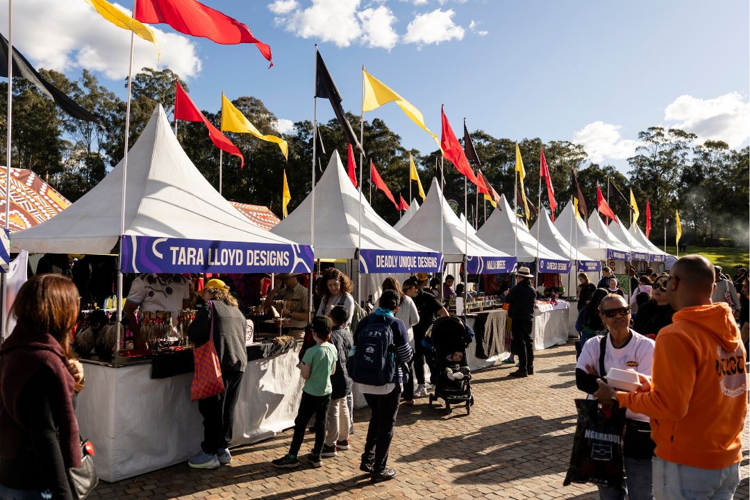 A lively outdoor market scene with white tents displaying colorful flags. People browse artisan stalls labeled "Tara Lloyd Designs" amid a clear blue sky.