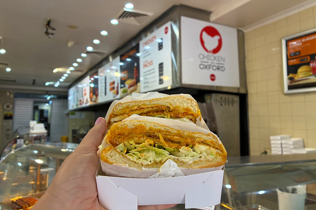 A hand holds a cut chicken sandwich with lettuce and sauce in a fast-food restaurant. The menu and "Chicken Oxford" sign are visible in the background.