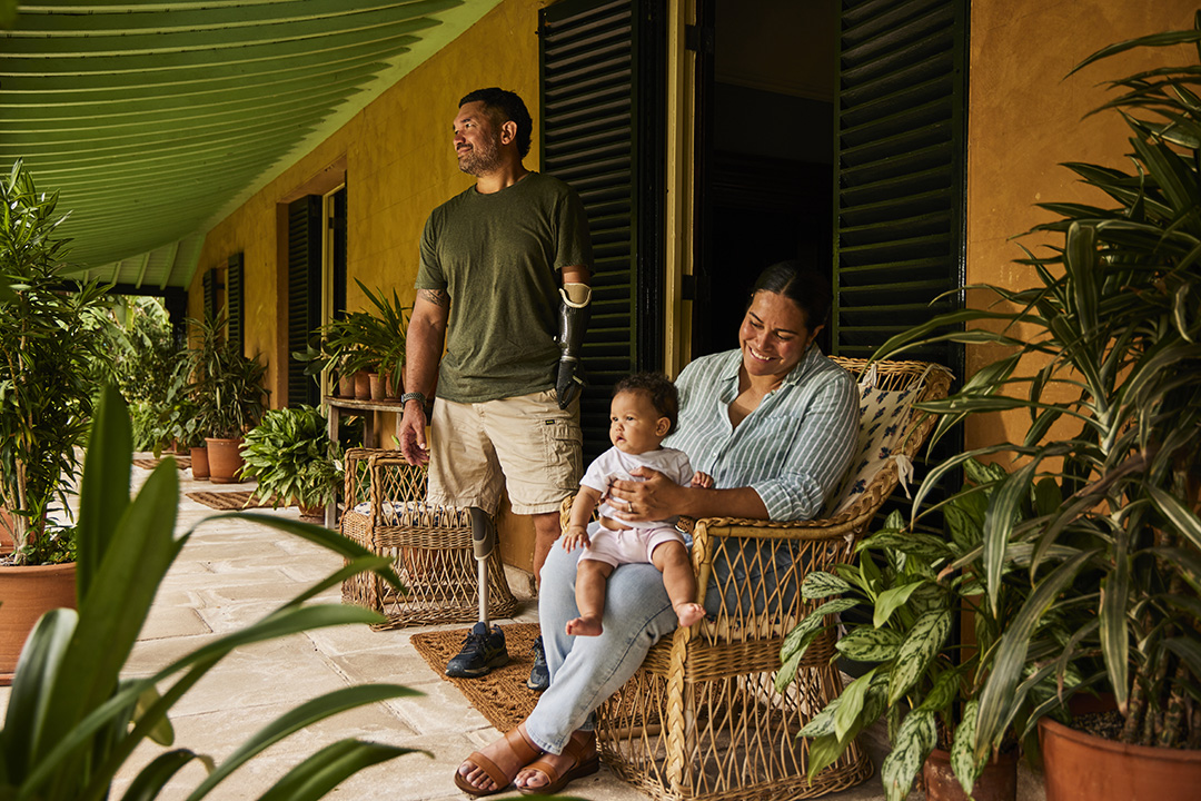 A man with a prosthetic leg and arm stands on a porch surrounded by plants, while a woman holding a baby sits nearby. The atmosphere is warm and relaxed.