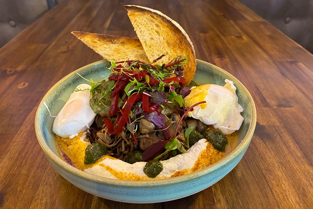 A vibrant dish with vegetables, greens, roasted meat, two poached eggs, and toast, garnished with herbs and sauce, on a wooden table.