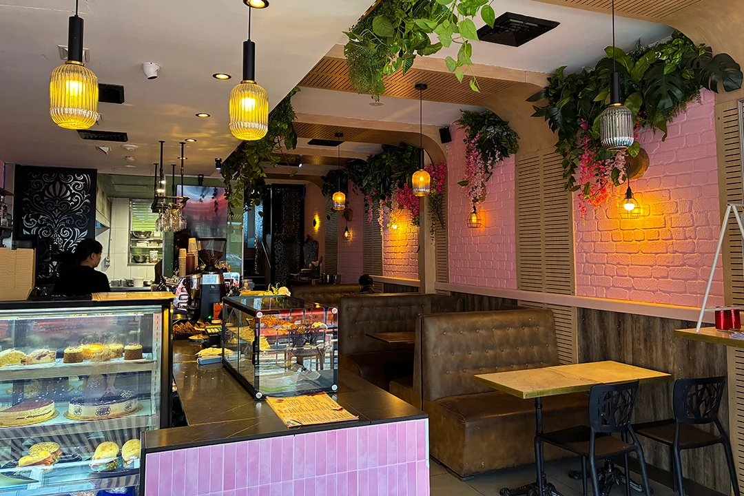 Cozy café interior with pink walls, greenery, and hanging lights. Display cases show pastries, while cushioned seating adds a warm atmosphere.