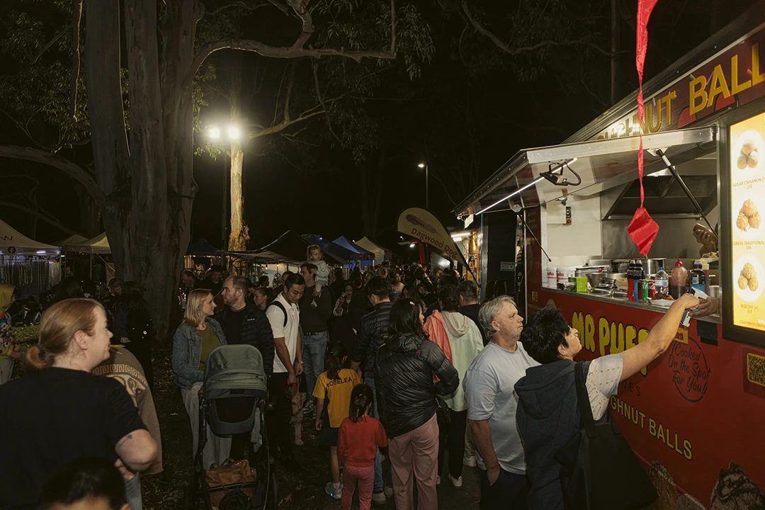 A bustling night market scene with a crowd gathered around a food truck selling doughnut balls. Bright lights illuminate the lively atmosphere.