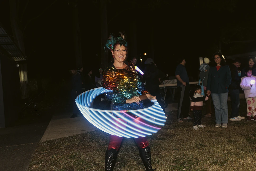 Woman hula hooping with glowing LED hoop at night, wearing a sparkling outfit. Onlookers watch, creating a festive, joyful atmosphere.