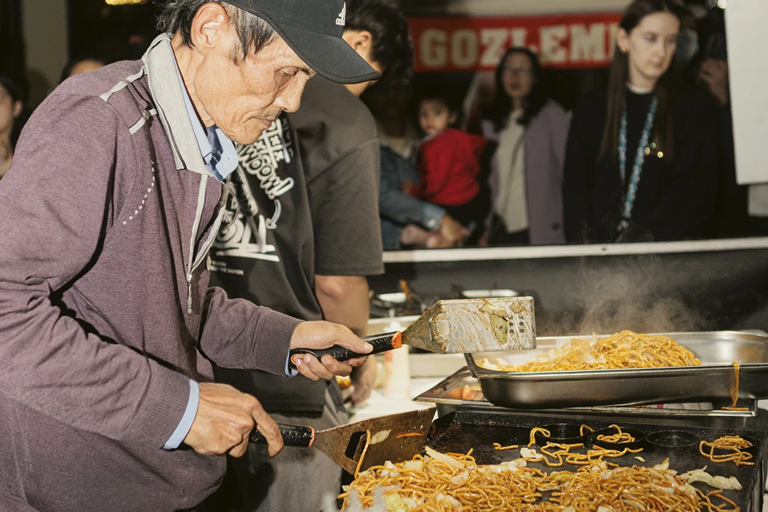 A man in a cap cooks noodles with a spatula on a crowded street food stall. Steam rises as onlookers, including a child, watch attentively.