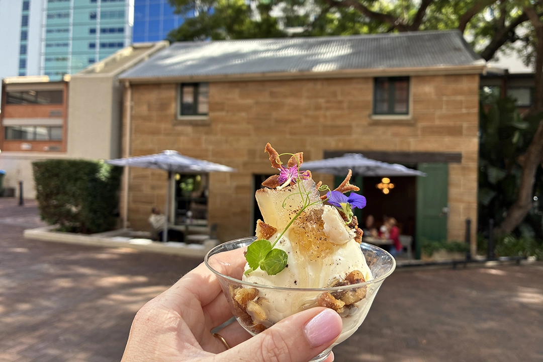 A hand holds a glass of ice cream topped with nuts and flowers, set against a rustic brick building under a blue sky, conveying a summery vibe.
