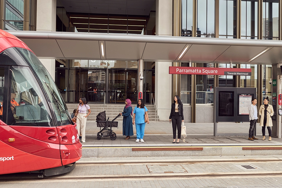 A red tram arrives at Parramatta Square station. Diverse individuals, including a woman with a stroller and a person in blue scrubs, wait on the platform.