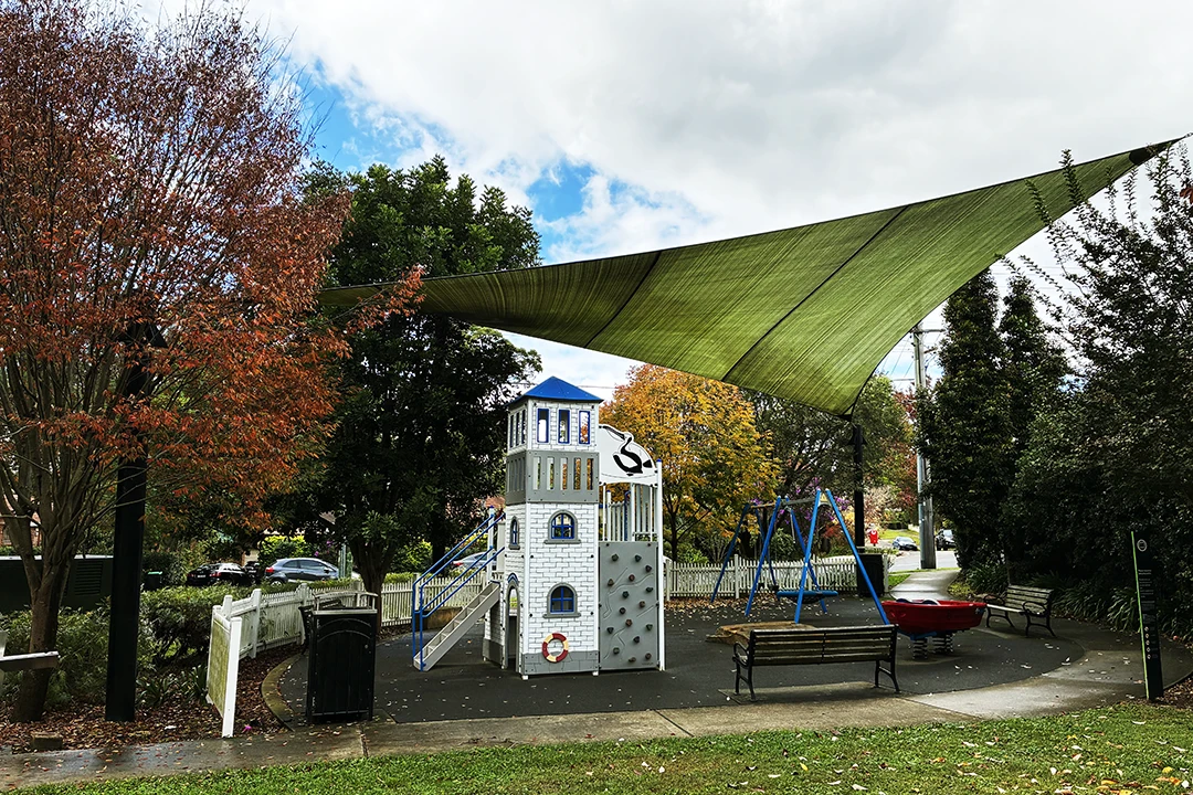 Playground with a white ship-themed climbing structure, swings, and red boat, shaded by a large canopy. Surrounding trees show autumn colours.