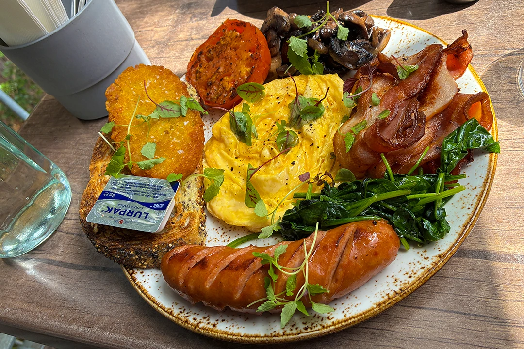 A vibrant breakfast plate on a wooden table with scrambled eggs, toast, bacon, sausage, spinach, mushrooms, grilled tomato, garnished with herbs.
