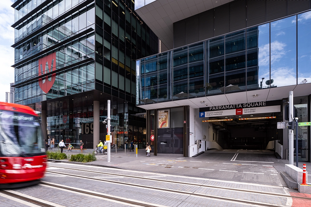 Modern urban streetscape featuring a glass building with a large red logo, pedestrians crossing, and a red tram passing by on tracks.