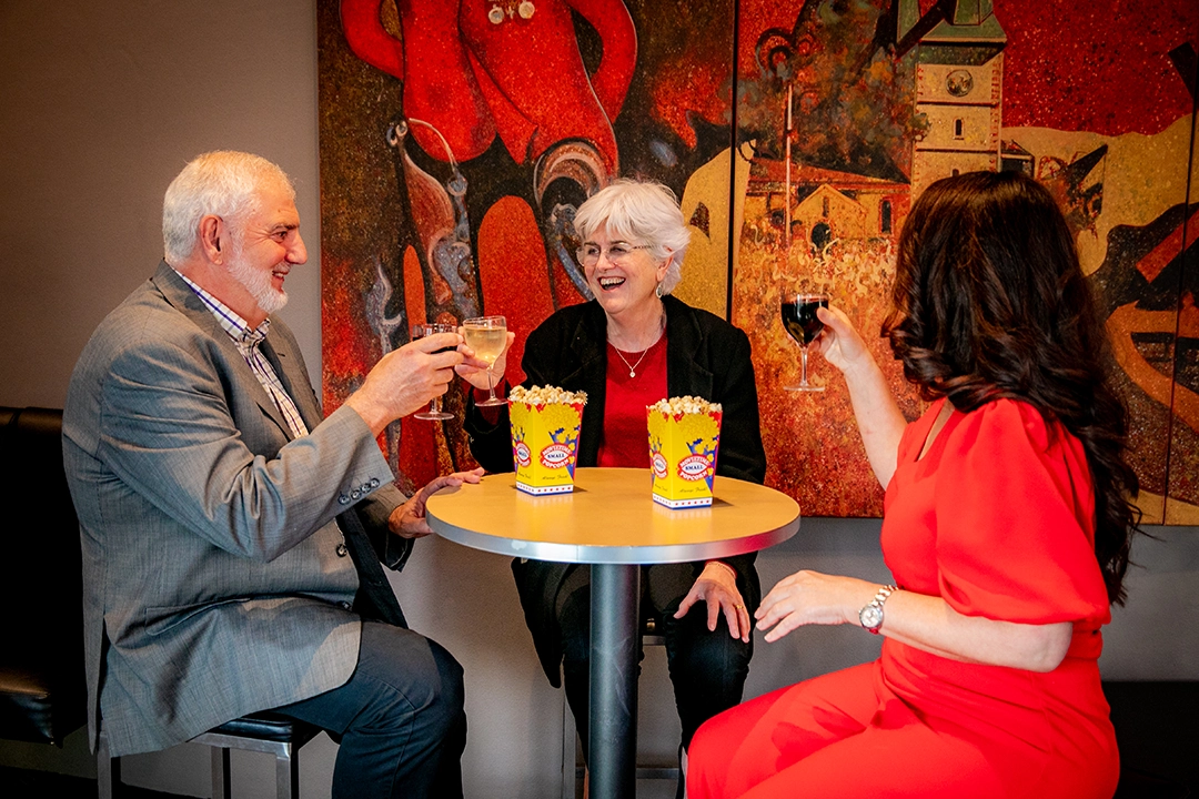Three people are seated around a small table, raising wine glasses in a toast. They are smiling and there are popcorn boxes on the table against a vibrant mural background.