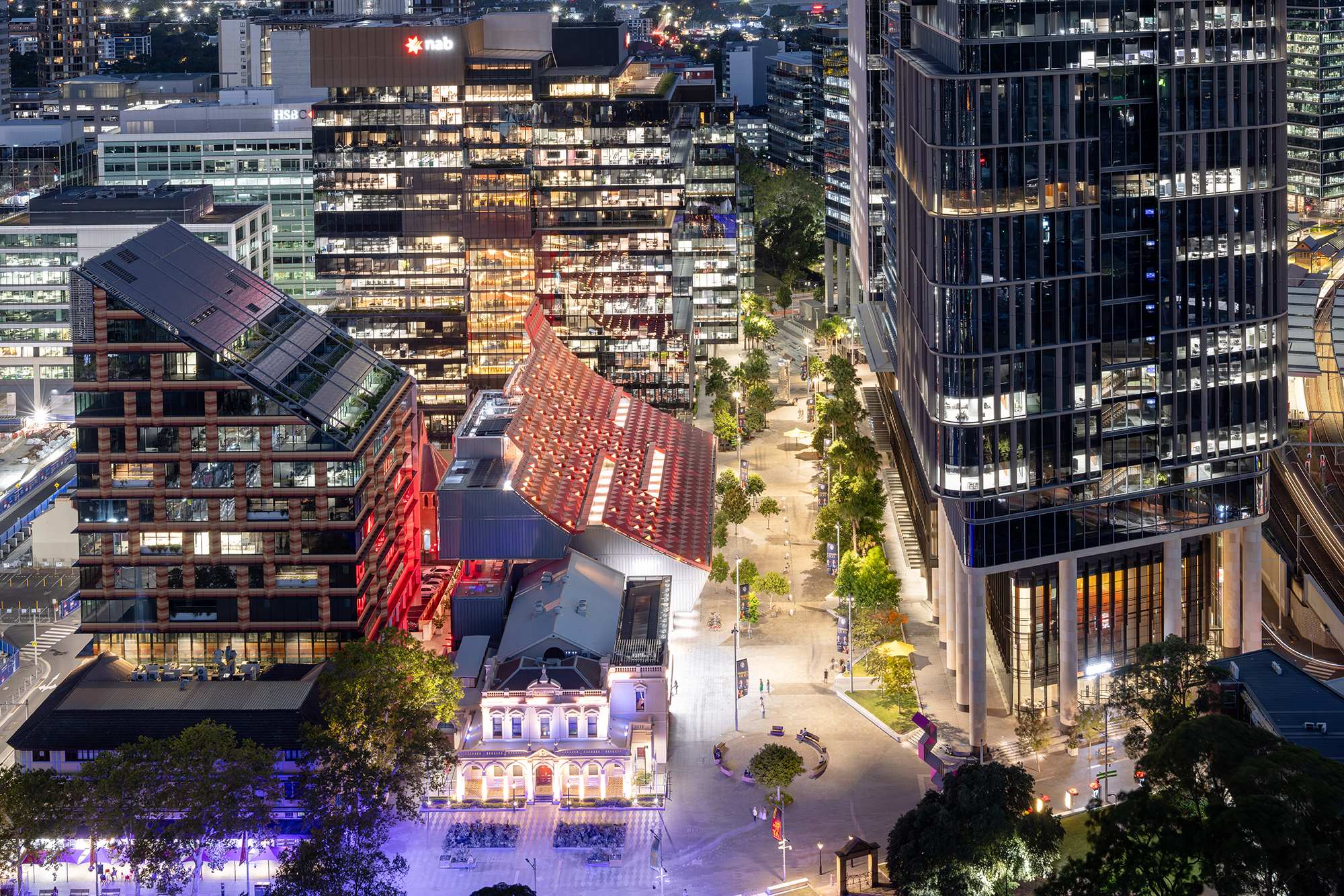 Aerial view of a vibrant cityscape at night, featuring illuminated modern skyscrapers, a curved-roof building, and a bustling tree-lined walkway.