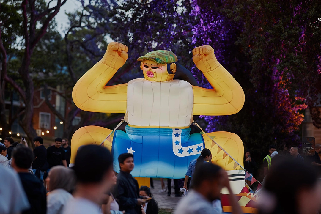 A large inflatable figure with yellow arms and a blue outfit is surrounded by a crowd at an outdoor event, set against a backdrop of trees and lights.