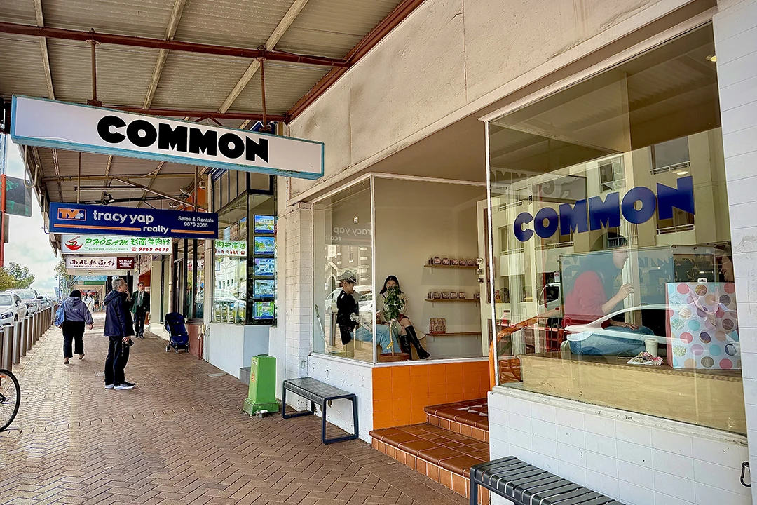 Street view of a shopfront with large windows labeled "COMMON" in bold letters. People walk by on a brick sidewalk, conveying a casual urban vibe.