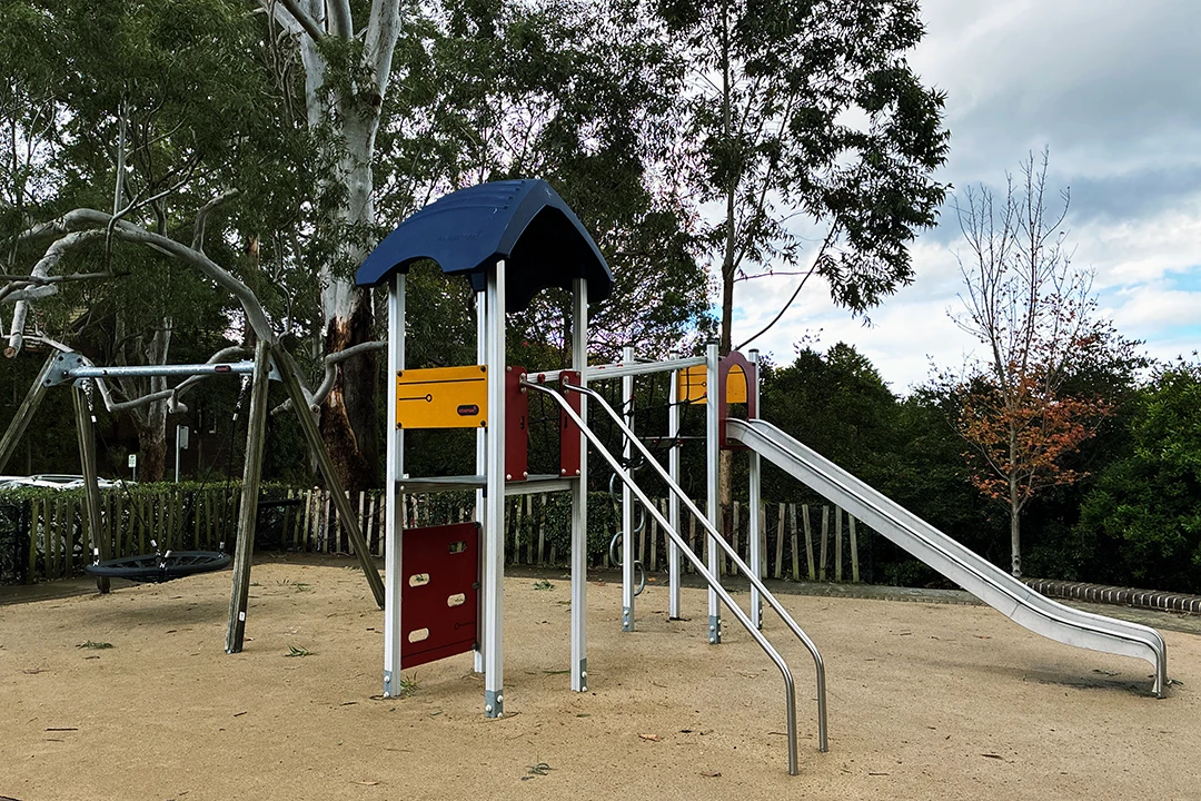 A colourful playground set features a slide, climbing structure, and swings, surrounded by trees under a partly cloudy sky, creating a calm atmosphere.