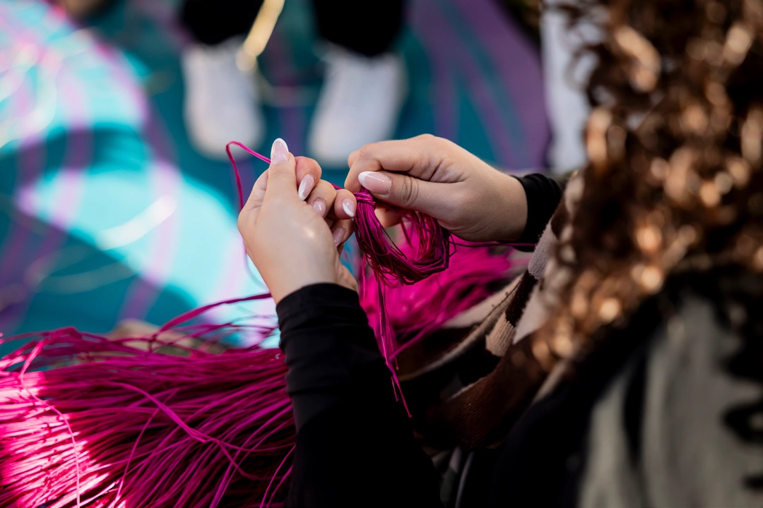 Hands delicately holding vibrant pink threads against a blurred, colorful background. The scene conveys focus and creativity.